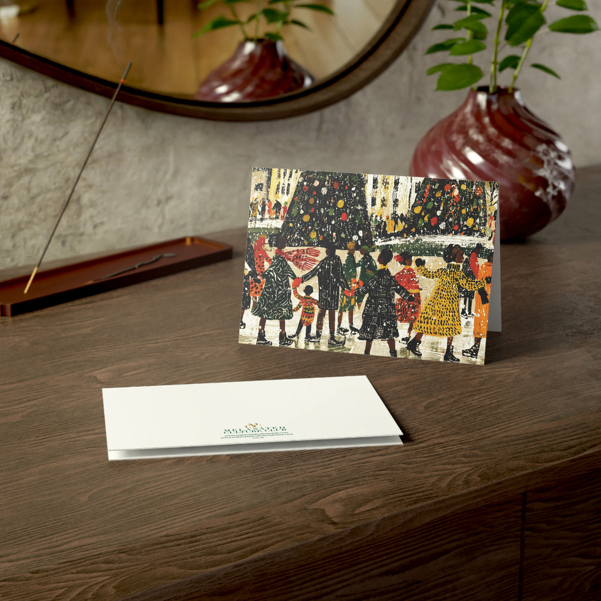 A Christmas greeting card and matching envelope displayed on a wooden windowsill, with a candle, plant, and eyeglasses nearby. The front of the card shows an illustrated winter scene with Black people skating, Christmas trees, and the back shows a small green brand logo.