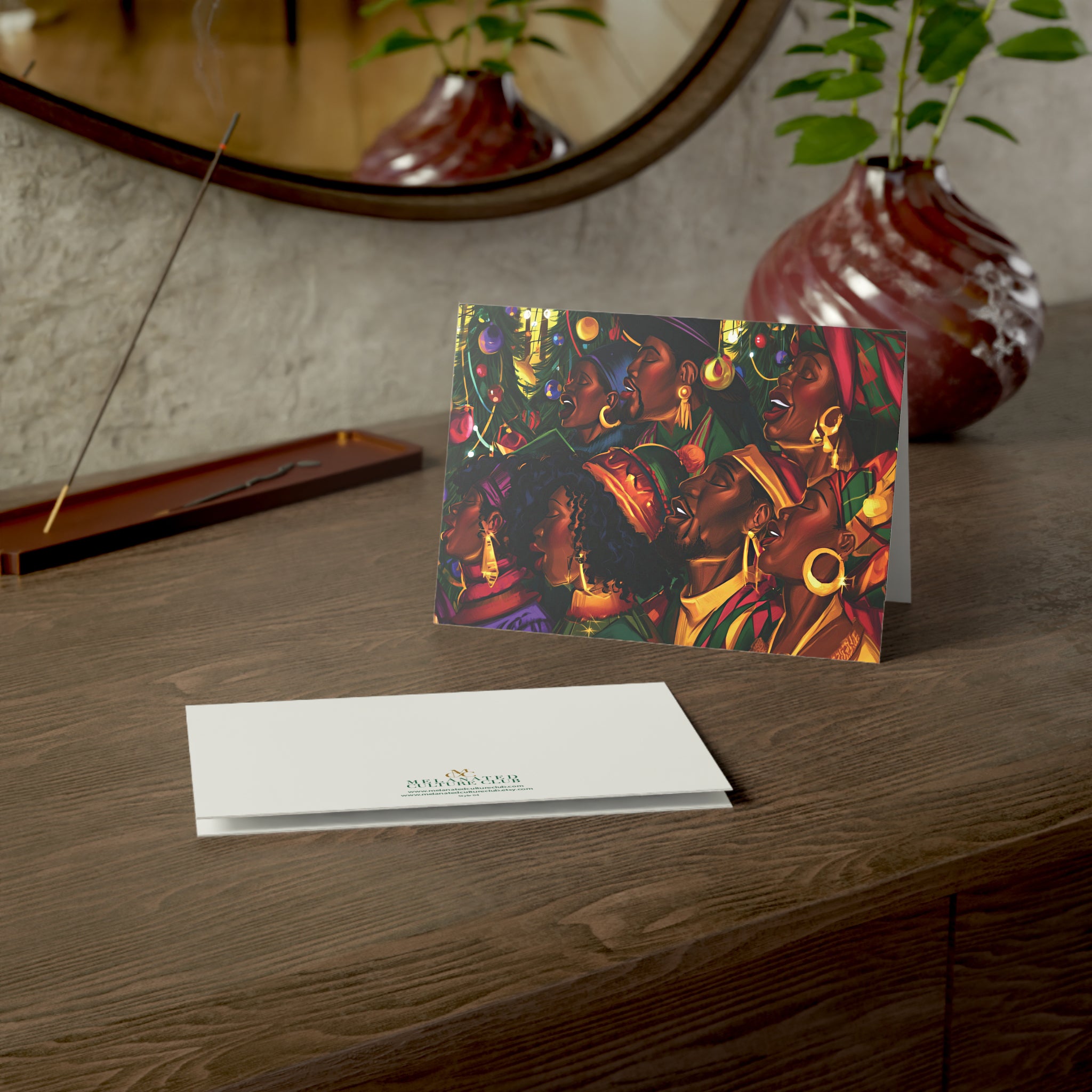 An Afrocentric holiday greeting card featuring a Black choir in black robes, , standing on a dark wooden table next to a white envelope printed with the brand logo. The background shows a textured wall, a mirror, and potted plants.