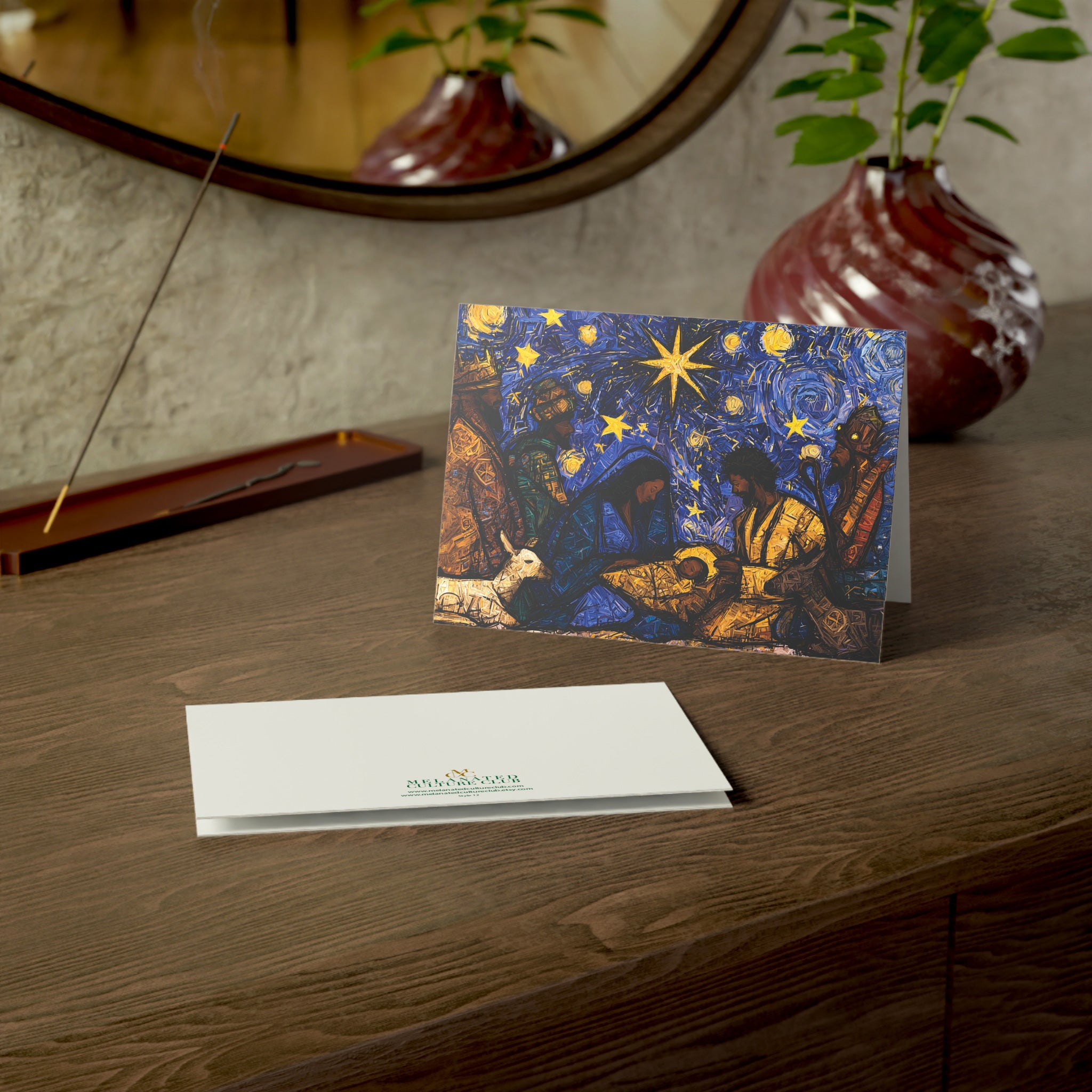 A Christmas greeting card featuring a colorful, stylized nativity scene with Black Baby Jesus, Mary, Joseph, Wise men and barn animals with a bright star in the night sky. A potted plant and a window are visible in the softly blurred background.
