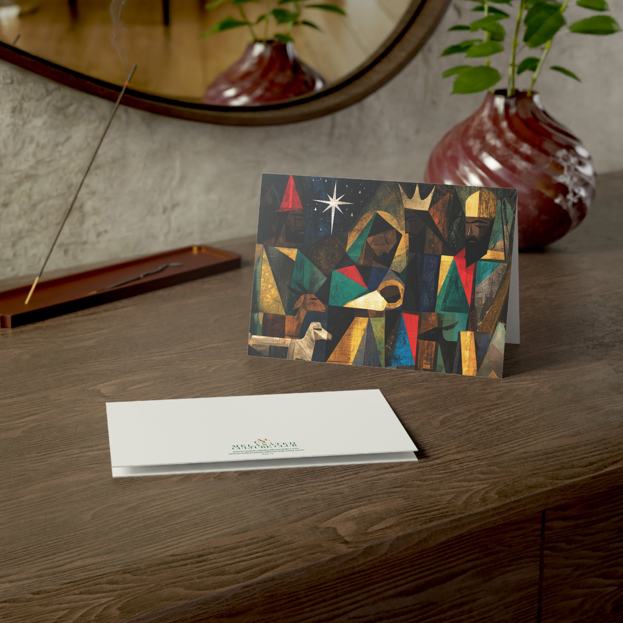 A Christmas greeting card featuring a colorful, stylized nativity scene with Black Baby Jesus, Mary, Joseph, Wise men and barn animals with a bright star in the night sky, on top of a wood surface.