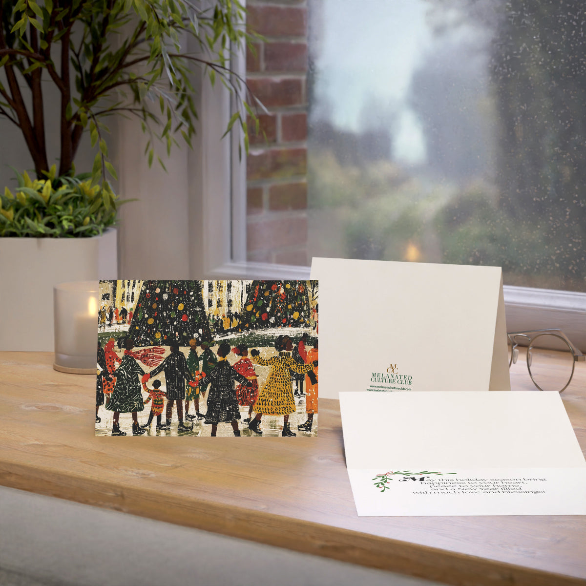 A Christmas greeting card and matching envelope displayed on a wooden windowsill, with a candle, plant, and eyeglasses nearby. The front of the card shows an illustrated winter scene with Black people skating, Christmas trees, and the back shows a small green brand logo.
