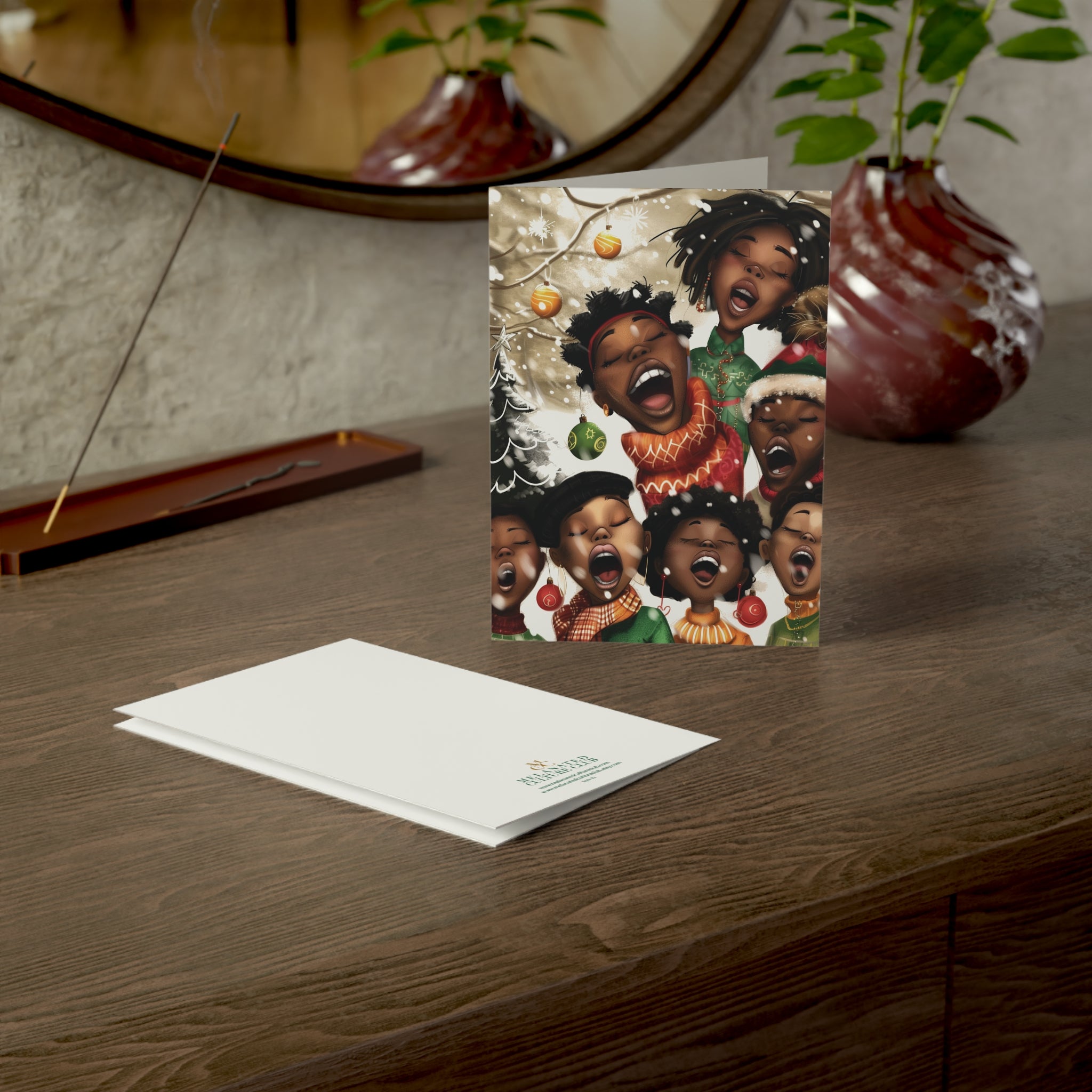 A Christmas-themed art print card featuring a group of Black children singing Christmas carols, displayed on a wooden table next to its white envelope. The background shows a warm, styled interior with a mirror, plants, and a vase.