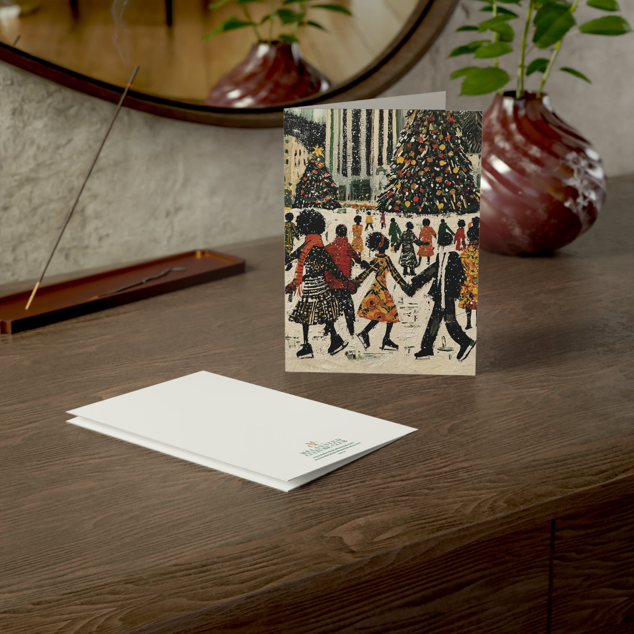 A square greeting card with a vintage-style illustration of Black people ice skating with Christmas trees in the background at the Rockefeller Center, stands on a wooden table, next to a folded white card with a small printed brand logo.