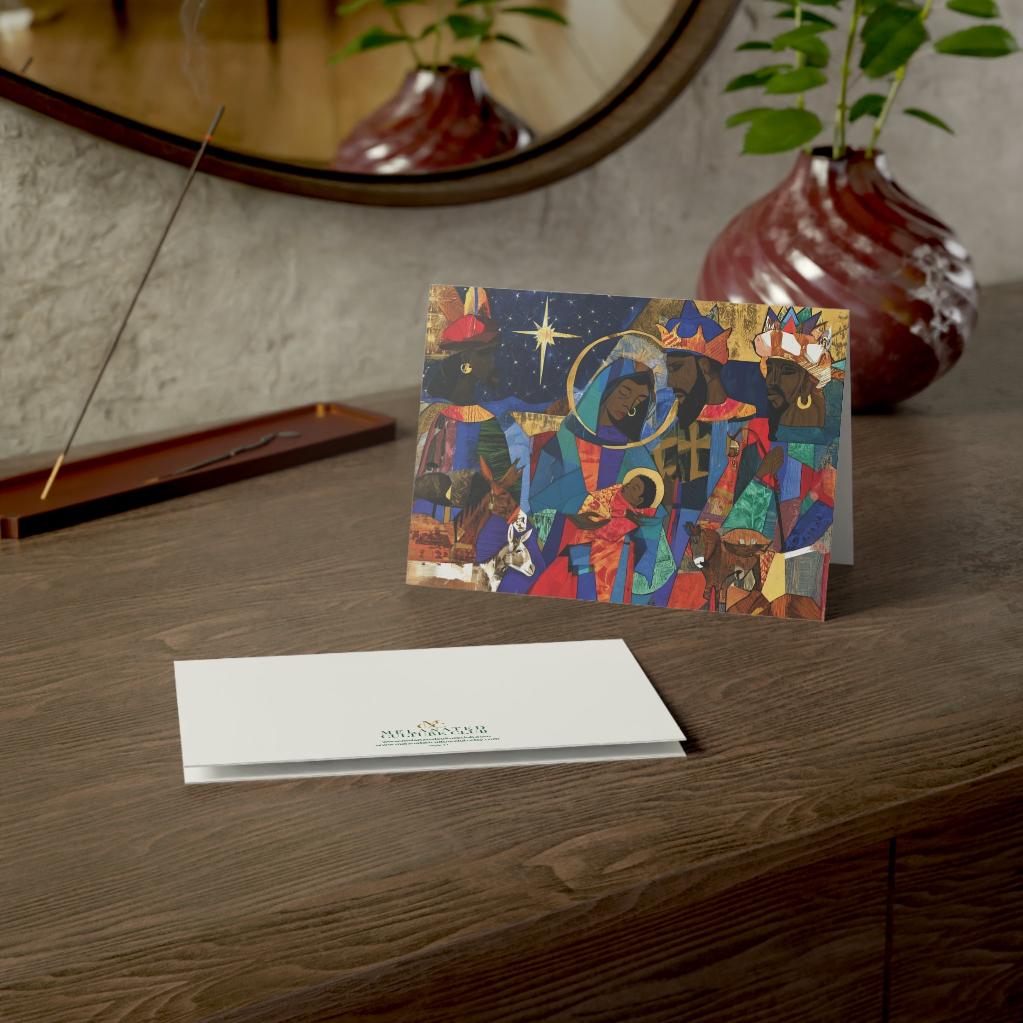 A Christmas greeting card featuring a colorful, stylized nativity scene with Black Jesus, Mary, Joseph, Wise men and barn animals with a bright star in the night sky, on top of a wood surface.
