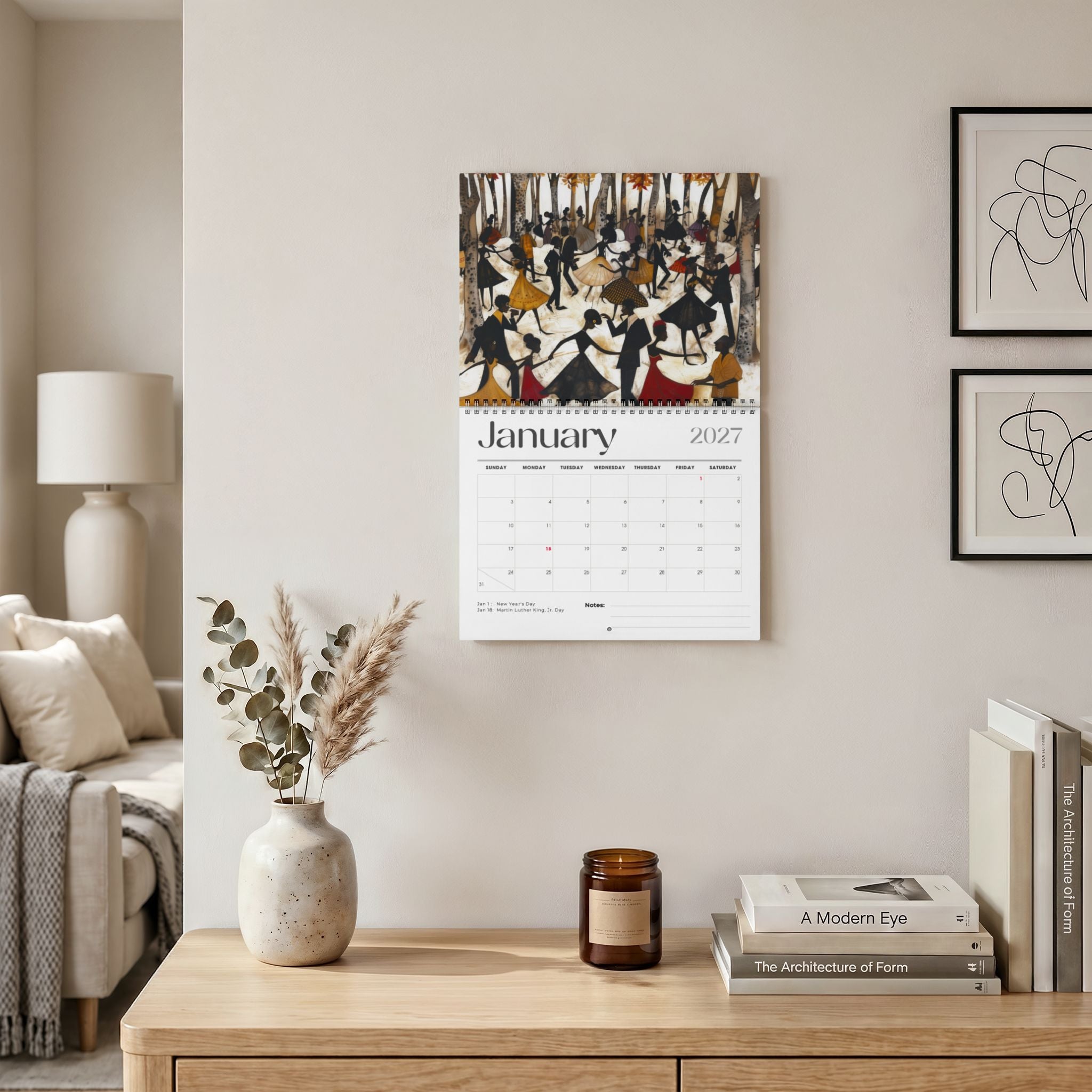 The Picnic in the Park 2027 wall calendar, featuring the month of January with abstract Black people dancing in celebration. Displayed on a beige wall above a wooden console table with a vase, candle, and books. Size 11" x 8.5"