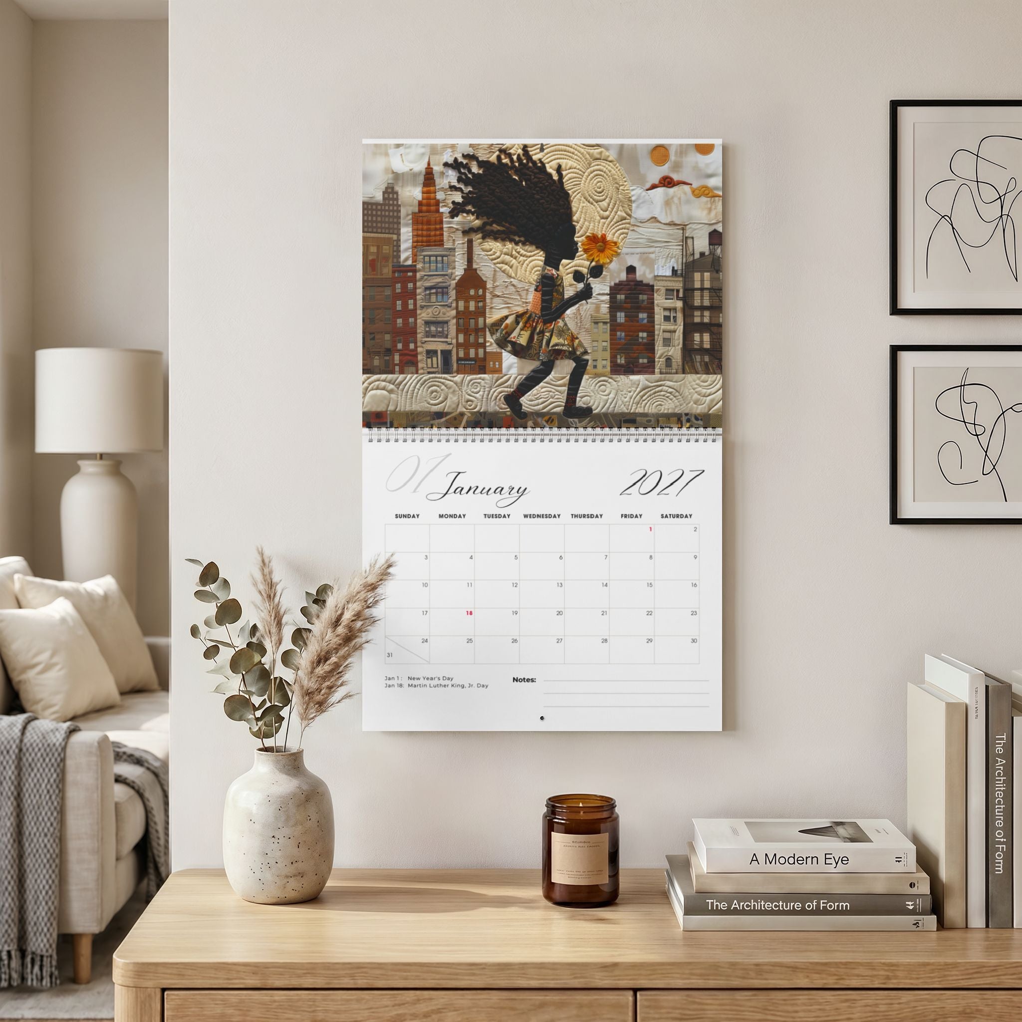 The Rooted in Home 2027 wall-mounted calendar hangs above a wooden console table in a styled living room. Size version 14" x 11.5".