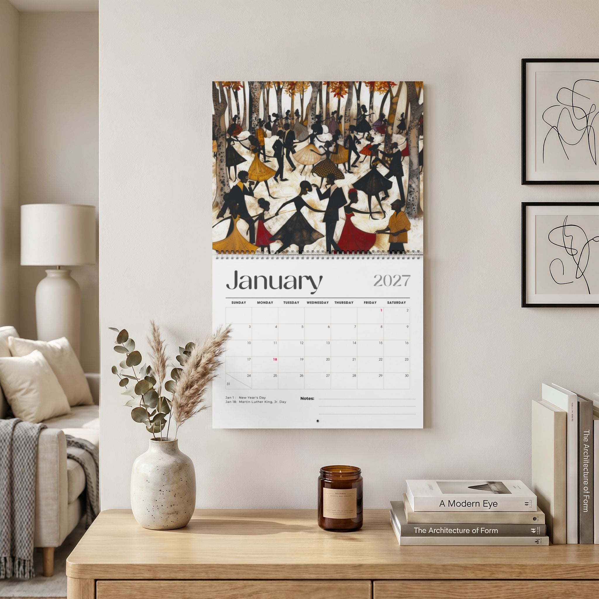 The Picnic in the Park 2027 wall calendar, featuring the month of January with abstract Black people dancing in celebration. Displayed on a beige wall above a wooden console table with a vase, candle, and books. Shown the 14" x 11" size calendar.