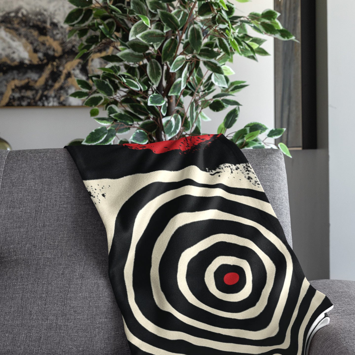 Folded view of Afrocentric geometric art throw blanket featuring bold black, red, gold, cream, and neutral abstract patterns with circles, spirals, grids, and textured African-inspired geometric shapes, draped over the end of a grey sofa.