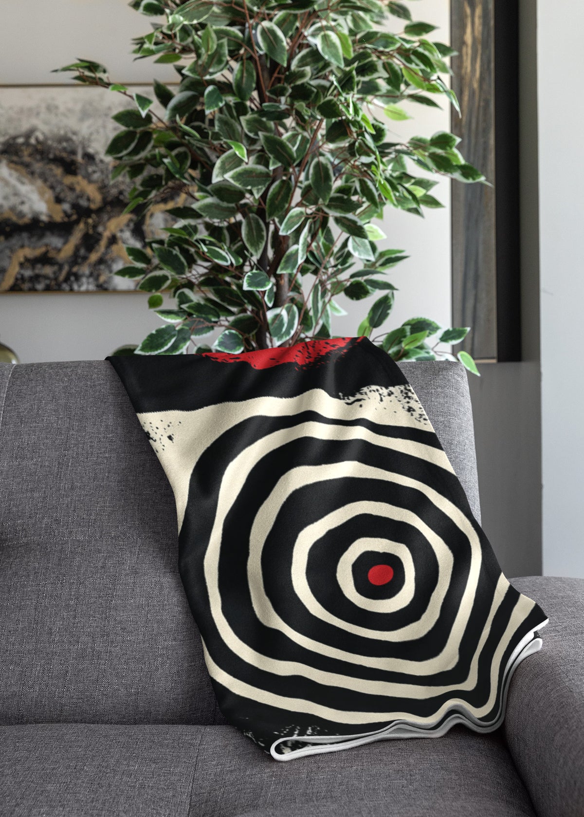 Folded view of Afrocentric geometric art throw blanket featuring bold black, red, gold, cream, and neutral abstract patterns with circles, spirals, grids, and textured African-inspired geometric shapes, draped over the end of a grey sofa.