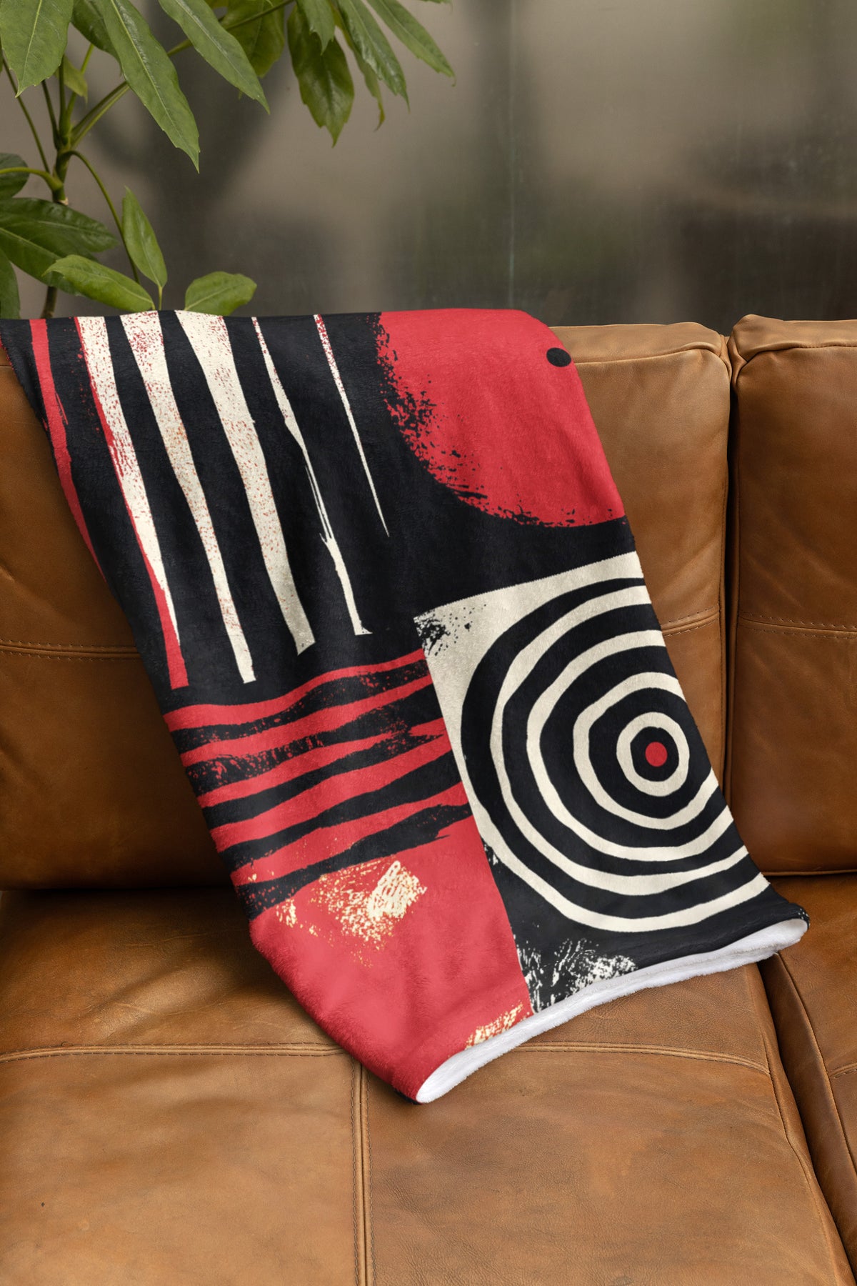 Folded view of Afrocentric geometric art throw blanket featuring bold black, red, gold, cream, and neutral abstract patterns with circles, spirals, grids, and textured African-inspired geometric shapes, draped over the back of a sable brown sofa.