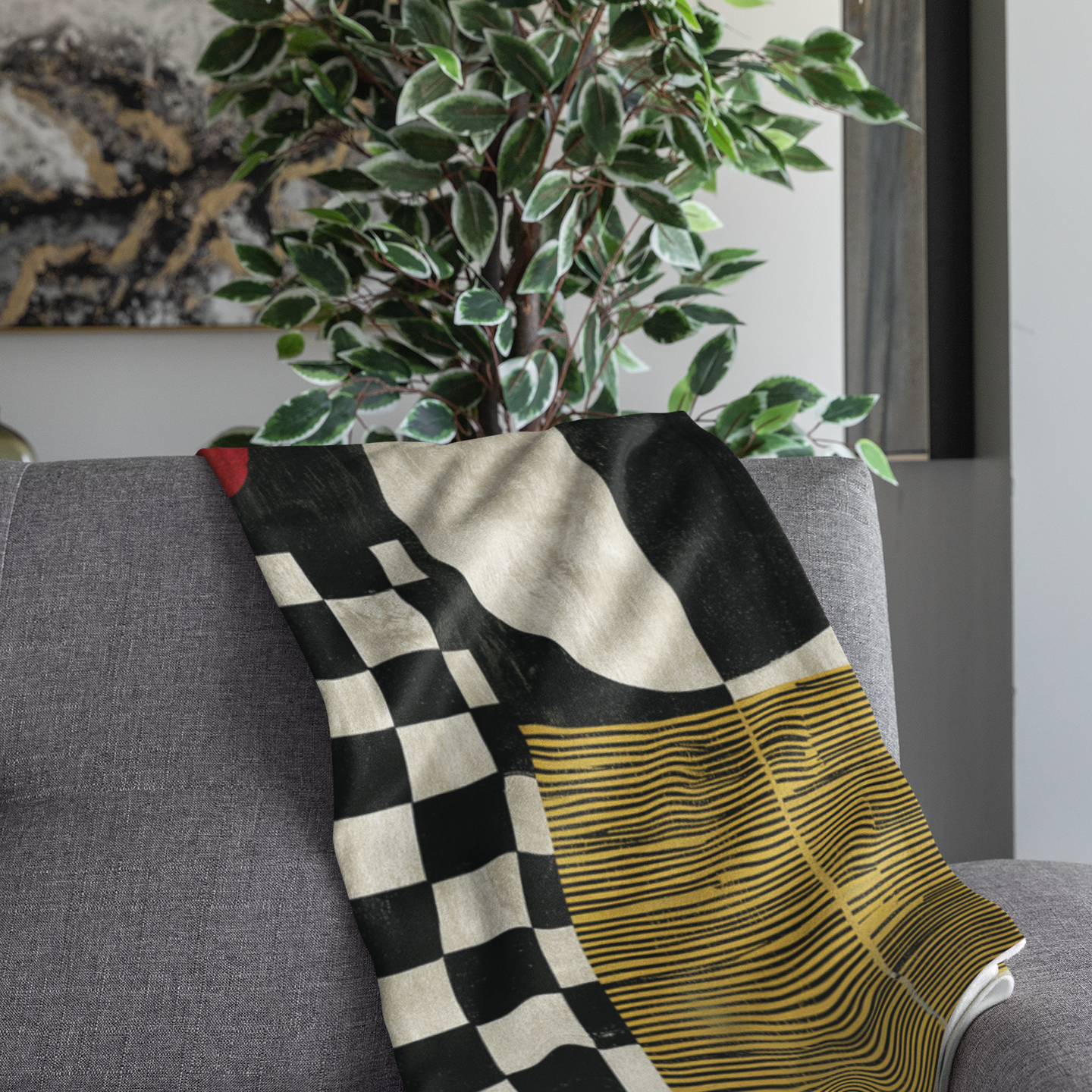 A folded throw blanket with a modern geometric pattern featuring a black and white checkerboard, a cream-colored curved section, and a yellow and black wavy striped section, draped over a gray sofa next to a potted plant.
