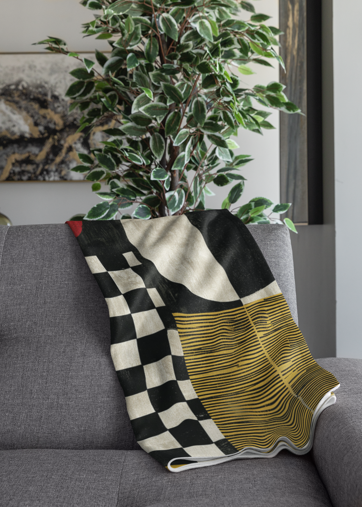 A folded throw blanket with a modern geometric pattern featuring a black and white checkerboard, a cream-colored curved section, and a yellow and black wavy striped section, draped over a gray sofa next to a potted plant.