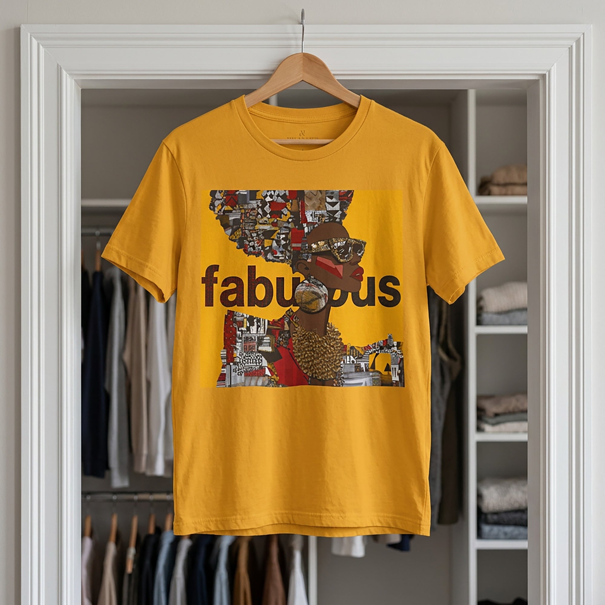 A mustard yellow short-sleeve t-shirt hanging on a wooden hanger in front of an open closet door. The shirt features an Afrocentric Black woman with a large graphic print on the front with the word 'fabulous' in dark brown text over a collage of colorful objects.