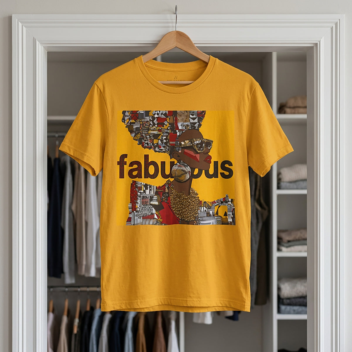 A mustard yellow short-sleeve t-shirt hanging on a wooden hanger in front of an open closet door. The shirt features an Afrocentric Black woman with a large graphic print on the front with the word 'fabulous' in dark brown text over a collage of colorful objects.
