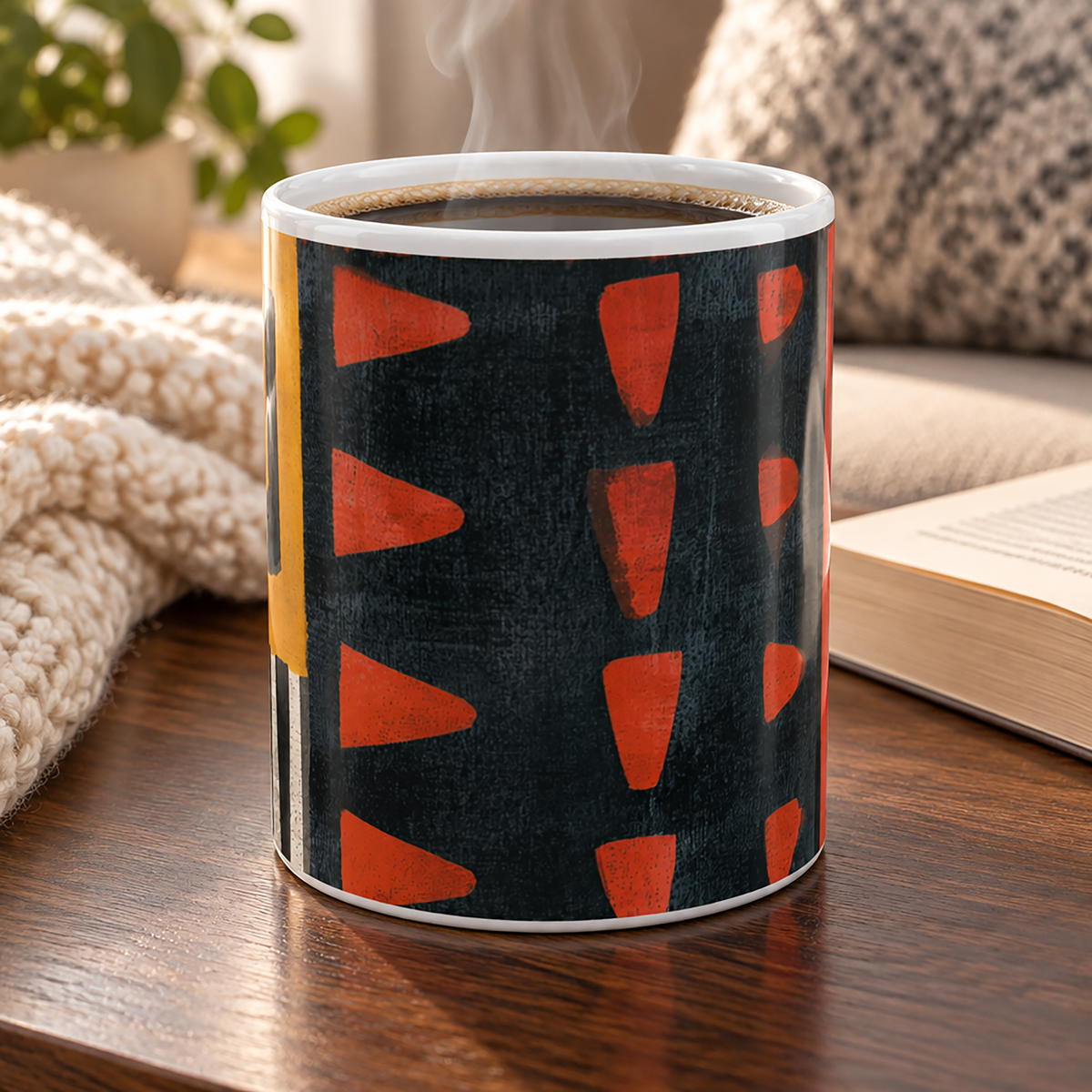 Front view of 11 oz Afrocentric abstract coffee mug featuring bold geometric shapes and textured patterns in red, gold, black, and cream, displayed on a wood table with open book and throw blankets.