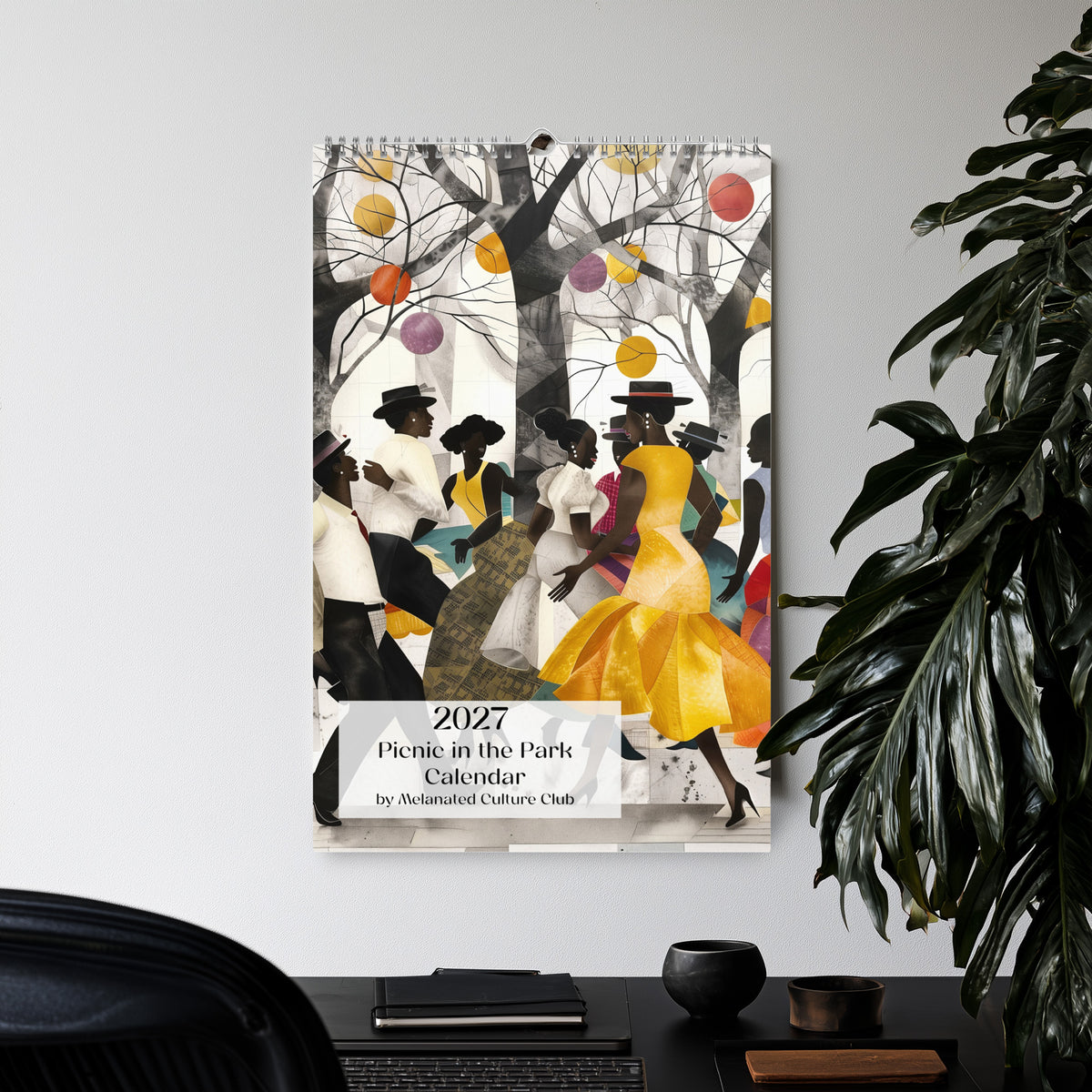 Front cover of an 11" x 17" 2027 Picnic in the Park Wall Calendar featuring vibrant African American artwork celebrating Black families enjoying a joyful outdoor picnic in a beautiful park setting. Colorful Afrocentric illustrations capture family gatherings, community, togetherness, and Black cultural traditions.  Displayed hanging on a wall in an office setting with a large green plant.
