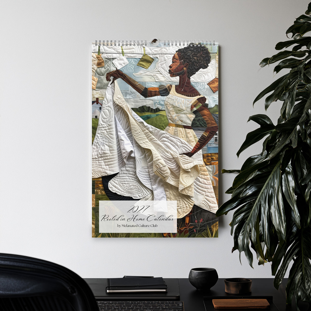 Displayed hanging on a wall in a office setting with a large green plant, an 11" x 17" 2027 Rooted in Home Wall Calendar featuring vibrant African American quilt collage artwork celebrating the everyday lives of Black women.