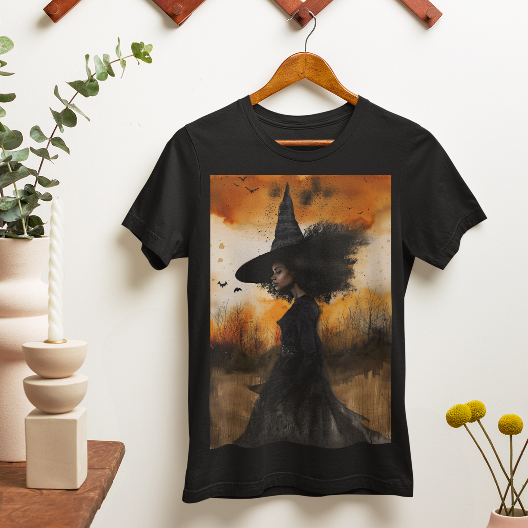 Afrocentric Black Witch T-Shirt featuring a watercolor illustration of an African American witch with black bats flying across a dark moody background. Unisex Halloween graphic tee with Afrocentric gothic design.
