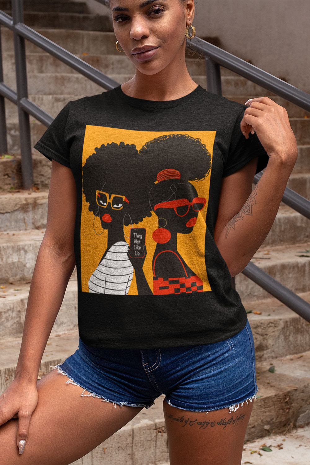 They Not Like Us Tee Shirt featuring two Black women in silhouette wearing high-fashion eyewear, holding a cell phone with the phrase "They Not Like Us" on an orange background – Afrocentric unisex apparel.