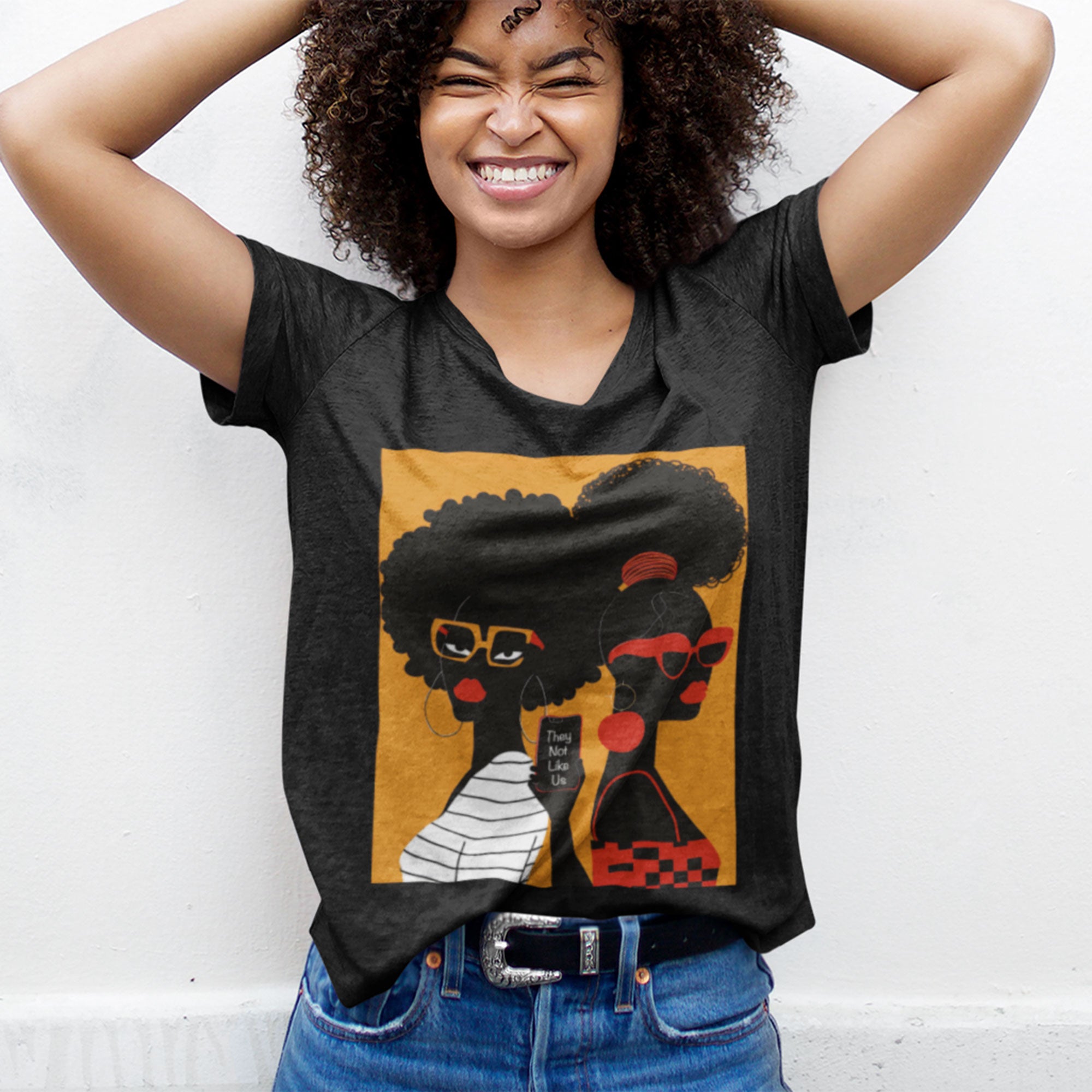 They Not Like Us Tee Shirt featuring two Black women in silhouette wearing high-fashion eyewear, holding a cell phone with the phrase "They Not Like Us" on an orange background – Afrocentric unisex apparel.