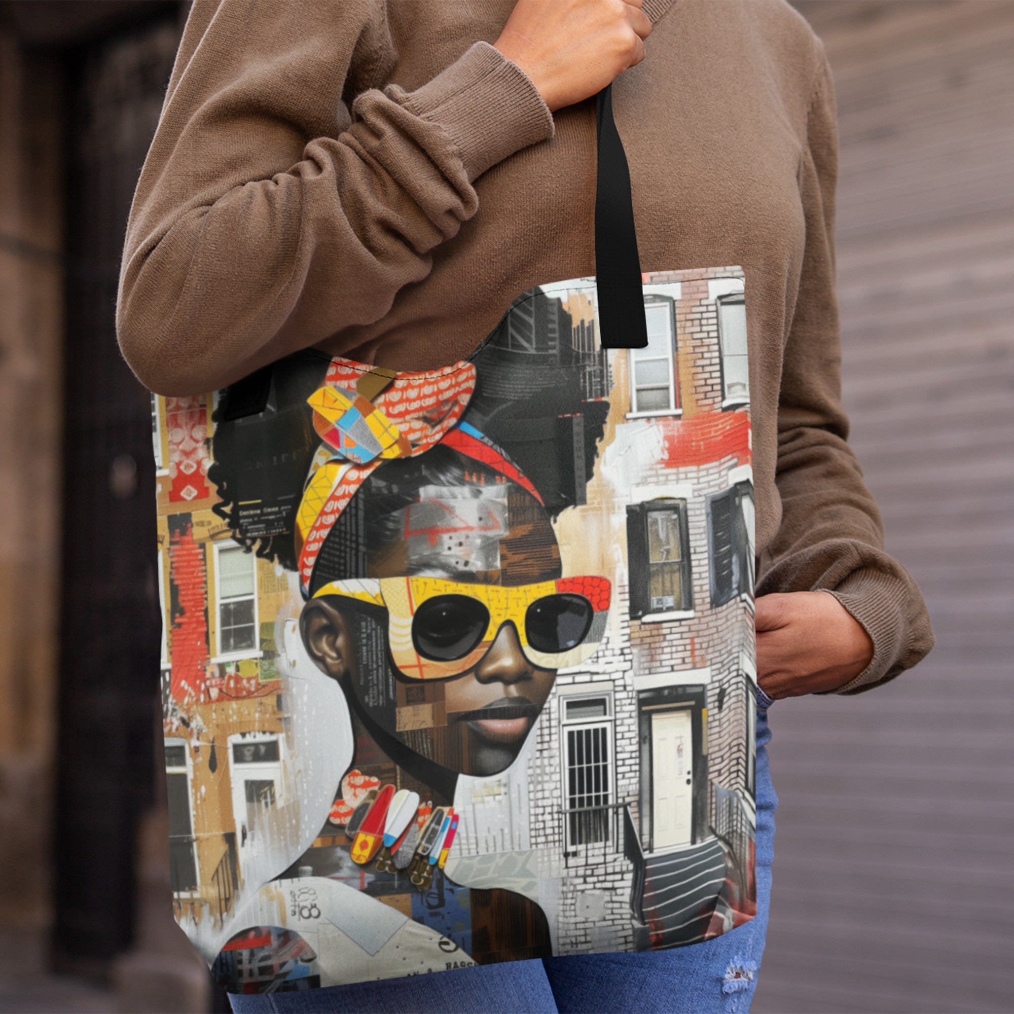 A black woman wearing a brown sweatshirt and blue jeans carries a large tote bag featuring a detailed collage-style portrait of a young black woman in sunglasses with colorful buildings in the background.