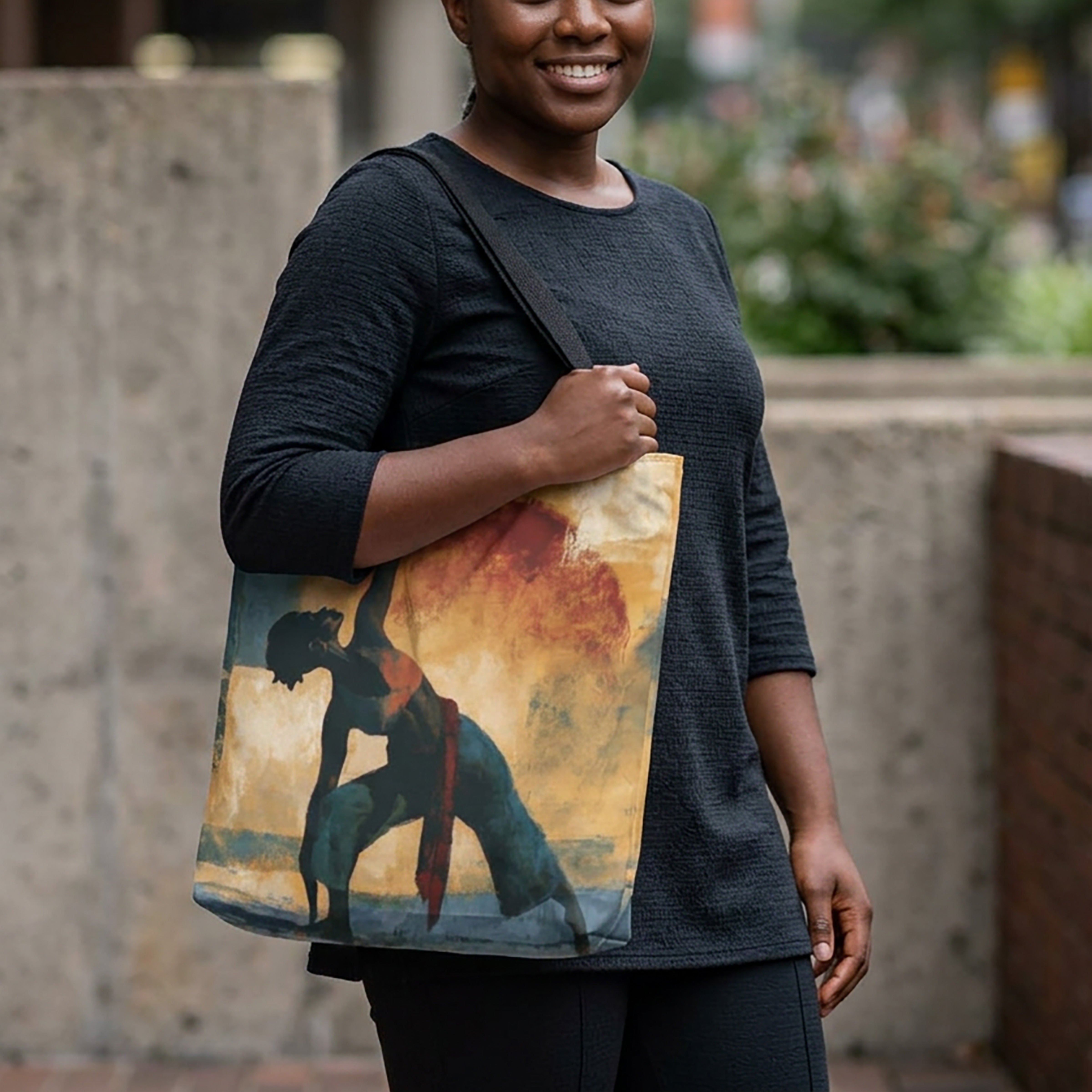 A Black woman wearing a dark charcoal gray knit top and black pants carries a canvas tote bag with a printed graphic of a black woman in a yoga pose against a warm sunset sky. The background is a softly blurred outdoor urban setting.