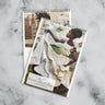 Views of front cover and the month of January of an 8" x 11" 2027 Rooted in Home Wall Calendar featuring vibrant African American quilt collage artwork celebrating the everyday lives of Black women, displayed against a grey marble background.